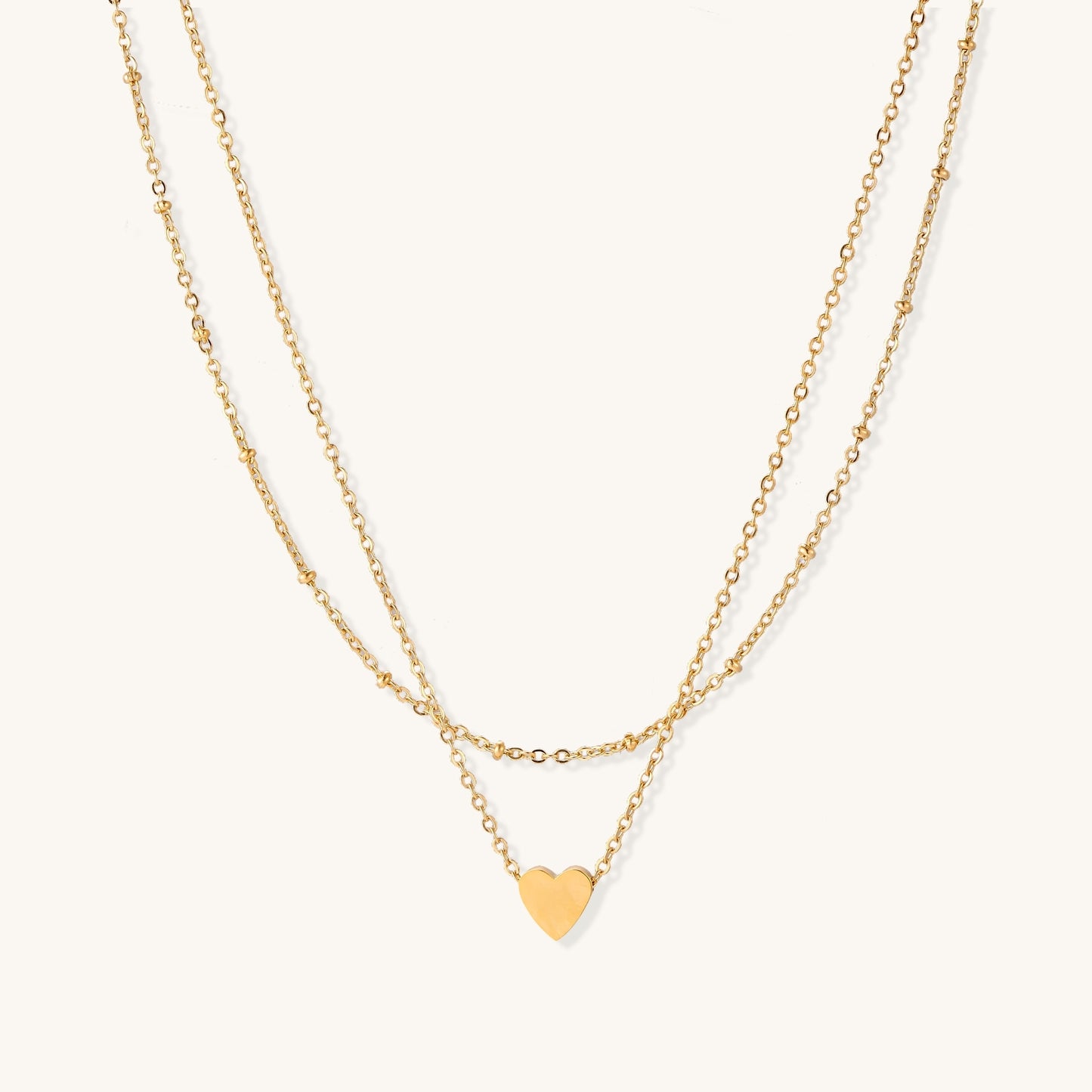 Two-layer gold-plated stainless steel necklace with small heart pendant displayed on beige background